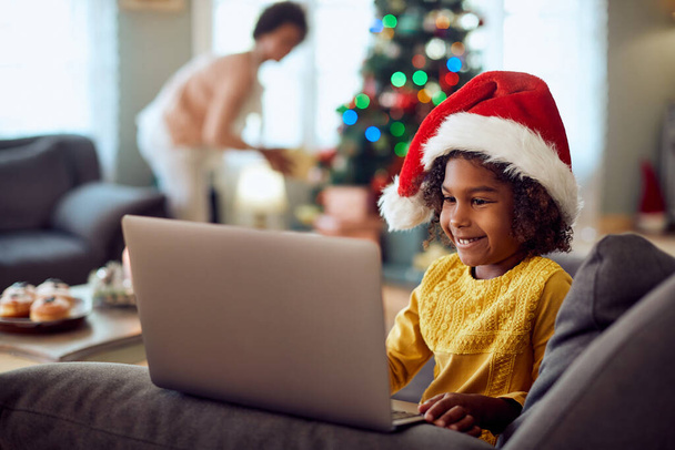 Happy African American girl wearing Santa hat and using laptop while relaxing at home for Christmas. Her mother is in the background. - Photo, Image
