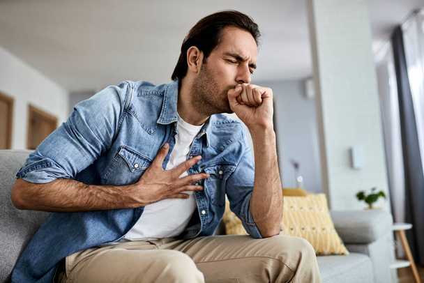 Sick man holding his chest in pain while coughing in the living room.  - Foto, afbeelding