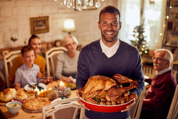 Happy man having traditional Thanksgiving meal with his family and holding roast turkey while looking at camera. - Foto, Imagem