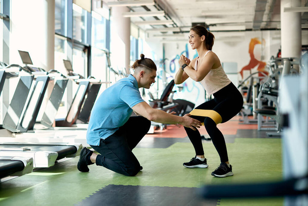Spor salonunda bacaklarında güç bandıyla çömelme egzersizleri yaparken atletik bir kadına asistanlık yapıyor.. - Fotoğraf, Görsel