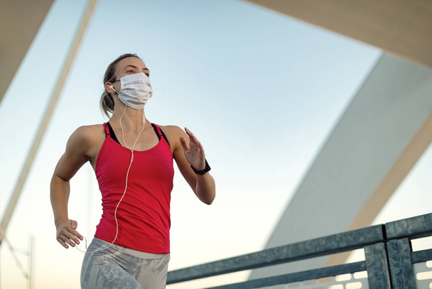 Low angle view of athletic woman wearing protective face mask while running outdoors during coronavirus pandemic. Copy space.  - Φωτογραφία, εικόνα
