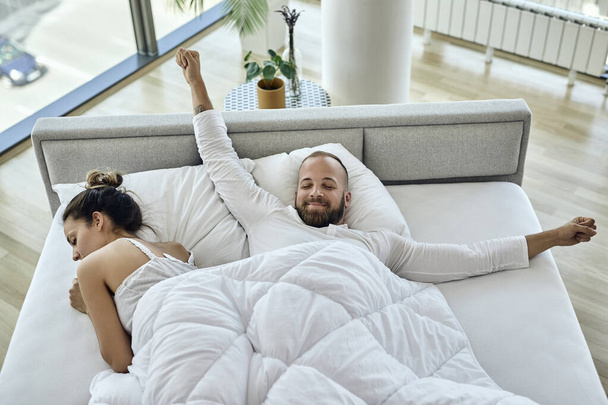 Високий кут зору щасливого чоловіка, що тягнеться вранці, лежачи в ліжку зі своєю дівчиною
.  - Фото, зображення