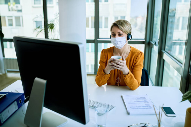 Businesswoman wearing face mask while working on a computer and drinking coffee in the office.  - Foto, Bild