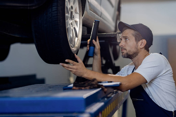 Közepes felnőtt szerelő autó gumiabroncsot vizsgál elemlámpával, miközben autószerelő műhelyben dolgozik.  - Fotó, kép