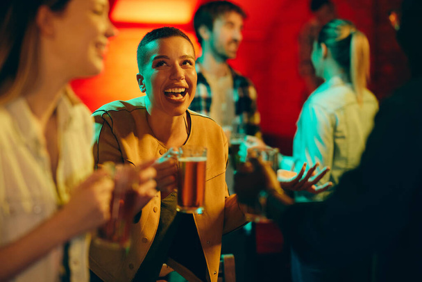 Young happy black woman drinking beer and having fun while being with her friends in a pub.  - Photo, Image