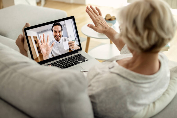 Close-up of mature woman waving while having video call with her son over laptop. Focus is on man on the screen.  - Photo, Image