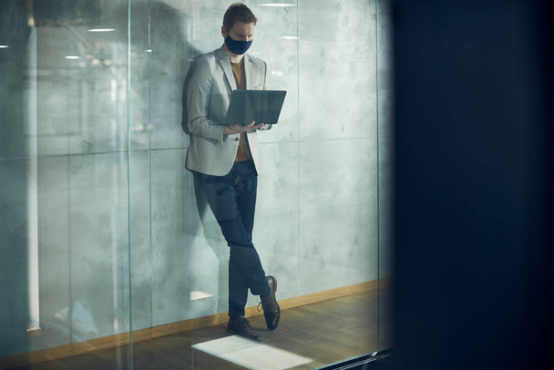 Male entrepreneur wearing face mask and working on laptop while standing in a corridor of an office building. The view is through the glass. Copy space. - Foto, imagen