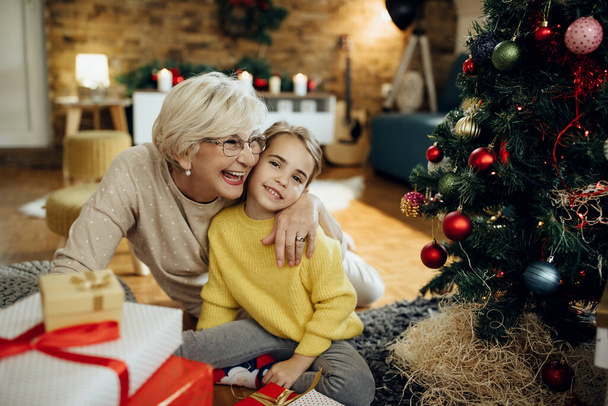 Boldog érett nő ölelgeti unokáját, miközben otthon pihen a karácsonyfa mellett..  - Fotó, kép