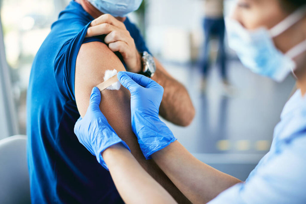 Nahaufnahme eines Gesundheitshelfers, der dem Patienten einen Klebeverband auf den Arm legt, nachdem er ihm im Impfzentrum den COVID-19-Impfstoff gegeben hat.  - Foto, Bild