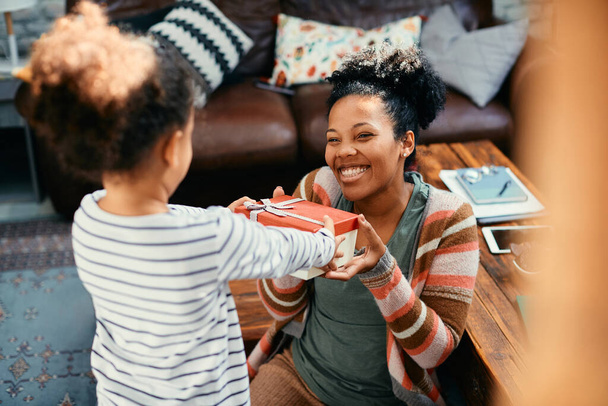 Afroamerykańska dziewczynka zaskakująca matkę pudełkiem prezentów w domu. Skup się na matce.. - Zdjęcie, obraz