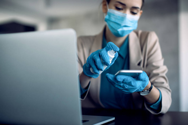 Close-up of businesswoman cleaning cell phone with disinfectant while working in the office during coronavirus pandemic.  - Photo, image