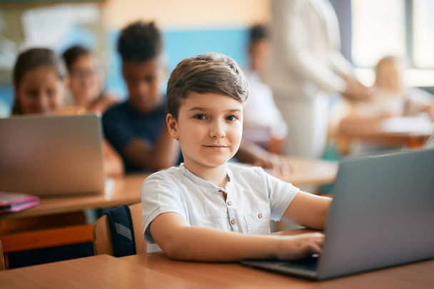 Милый школьник учится за ноутбуком во время занятий в классе и смотрит в камеру.  - Фото, изображение