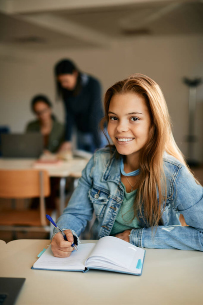 Счастливая девочка-подросток делает заметки во время учебы в школе и смотрит в камеру.  - Фото, изображение