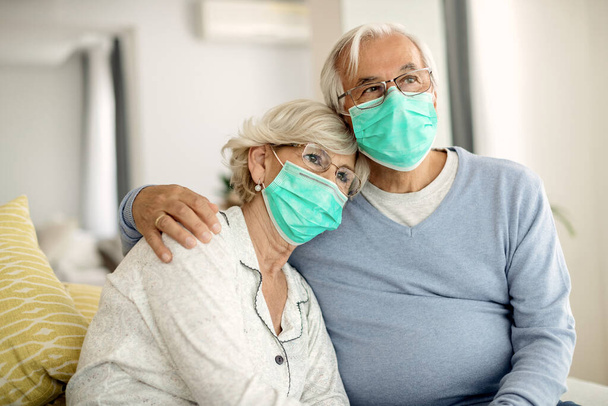 Pensive mature couple wearing protective face masks and feeling lonely during home quarantine due to coronavirus epidemic.  - Fotografie, Obrázek