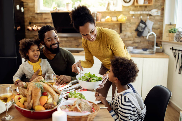 Happy African American woman having Thanksgiving lunch with her family and serving salad at dining table.  - Foto, imagen