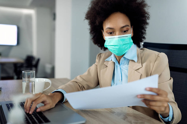 Black female entrepreneur using laptop and examining business reports while wearing face mask in the office.  - Foto, Bild