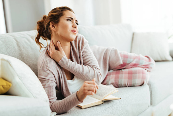 Young woman having neckache while lying down on sofa and reading book in the living room.  - Foto, afbeelding