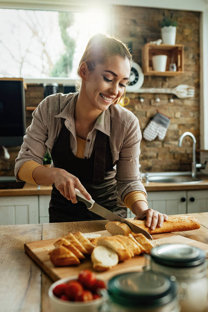 Young smiling woman slicing bread with a knife while preparing food in the kitchen.  - Photo, image