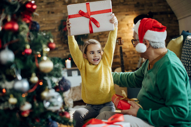 Щаслива маленька дівчинка отримує подарунок від діда і розважається під час Різдва вдома
.  - Фото, зображення
