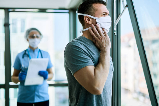 Pensive man with face mask feeling COVID-19 symptoms and looking through the window while being at medical clinic. Nurse is standing in the background.  - Фото, изображение