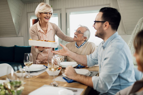 Happy mature woman serving lunch while talking to her family at dining table. - Foto, Bild