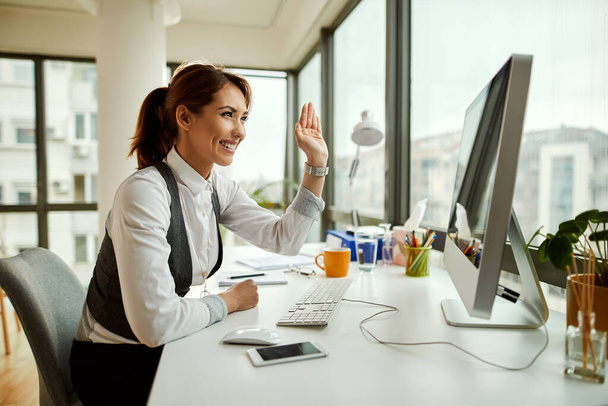 Молода щаслива бізнес-леді використовує комп'ютер і махає під час відеодзвінка в офісі
. - Фото, зображення