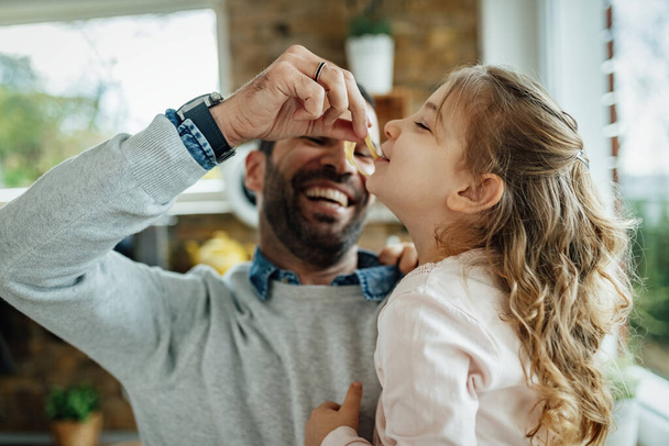Happy father having fun with his small daughter while feeding her in the kitchen. Focus is on little girl.  - Φωτογραφία, εικόνα