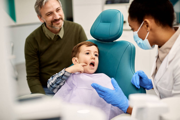 Маленький хлопчик розповідає про зубний біль у стоматолога під час перебування з батьком у стоматологічній клініці
.  - Фото, зображення