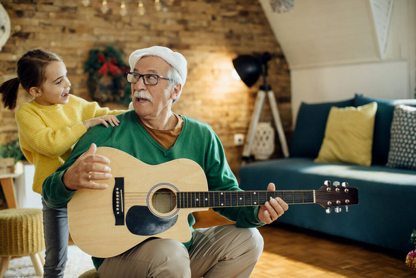 Šťastný senior muž hraje na akustickou kytaru, zatímco vnučka tančí kolem něj na Vánoce. - Fotografie, Obrázek