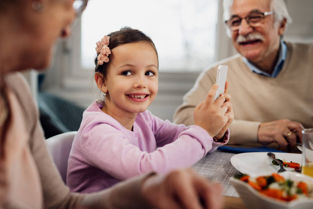 Happy little girl communicating with her grandparents while using cell phone during lunch at dining table.  - Foto, Imagem