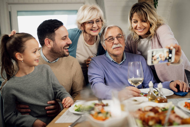 Счастливая семья из нескольких поколений, говорящая селфи с мобильным телефоном во время совместного обеда дома.  - Фото, изображение
