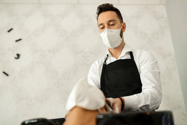 Низький кут зору перукаря в захисній масці для обличчя під час миття волосся жінки в салоні під час епідемії COVID-19
.  - Фото, зображення