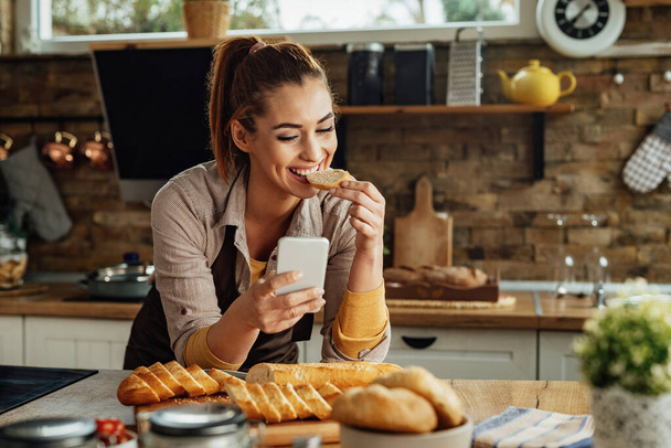 Young happy woman text messaging on mobile phone and eating bread in the kitchen.  - Foto, afbeelding