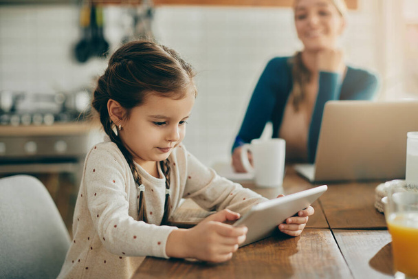 Little girl using touchpad and watching something on the Internet while her mother is working on computer in the background. - Foto, Imagen