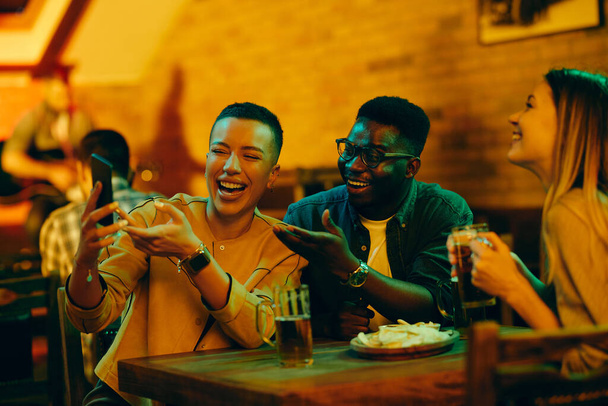 Ryhmä onnellisia ystäviä hauskaa juodessaan olutta ja käyttämällä matkapuhelinta baarissa.  - Valokuva, kuva