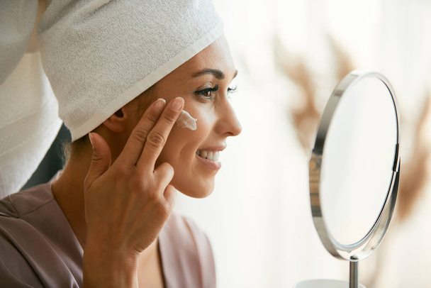 Young smiling woman using moisturizer while taking care of her face skin at home. - Foto, immagini