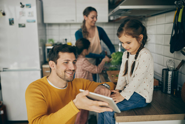 Happy father showing something to his daughter on digital tablet in the kitchen. Mother and other daughter are preparing food in the background.  - Foto, Imagem