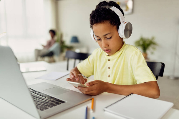 Small black boy wearing headphones and texting on mobile phone while learning at home. - Foto, Imagen