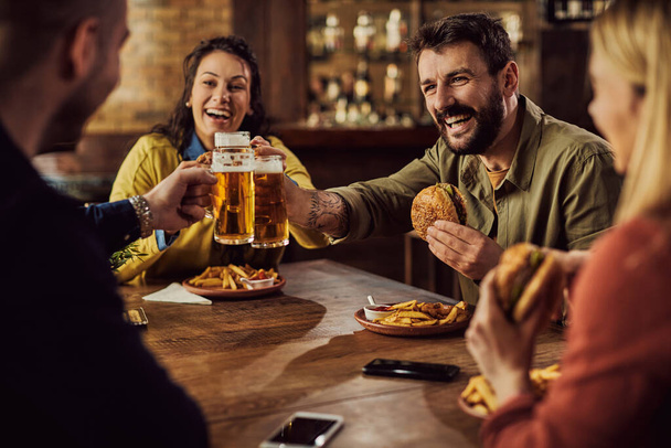 Cheerful man and his friends toasting with beer while eating hamburgers in a pub. - Foto, afbeelding
