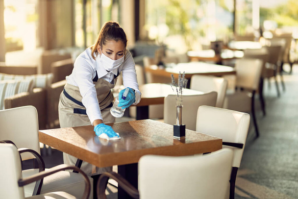 Молодая официантка дезинфицирует столы, надевая защитную маску для лица и перчатки из-за эпидемии коронавируса.  - Фото, изображение