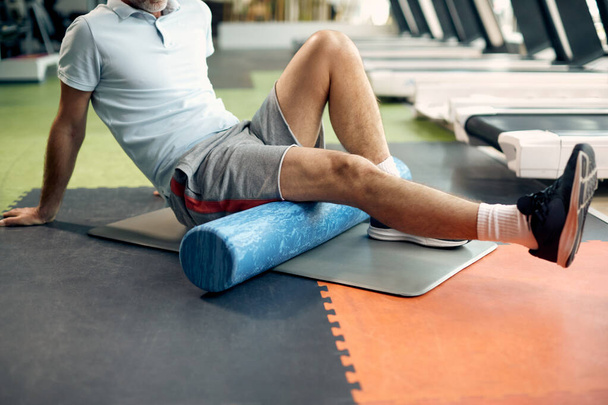 Onherkenbare volwassen atleet masseert zijn benen met schuimroller na sporttraining in een fitnessruimte.  - Foto, afbeelding