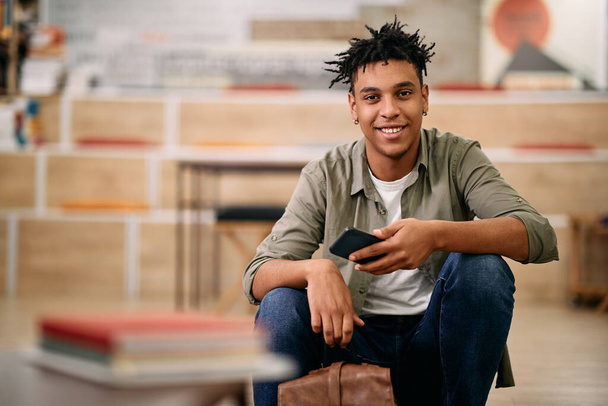 Portrait of young happy black student using mobile phone at university library looking at camera. - Foto, imagen