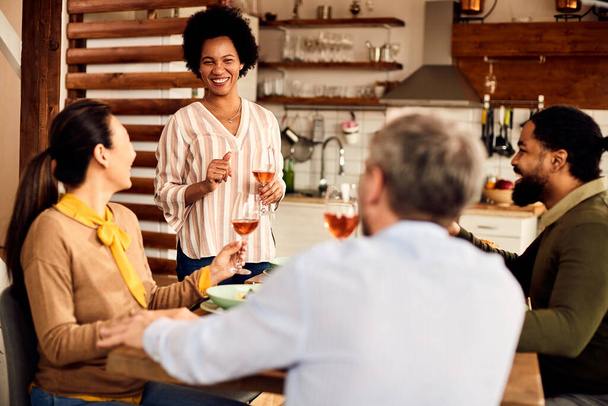 Multi-ethnic group of happy friends having lunch and drinking wine in dining room. Focus is o African American woman holding a toast.  - Foto, Imagen