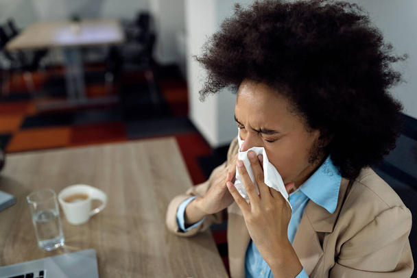 Black businesswoman blowing nose in a tissue while working in the office.  - Foto, Bild
