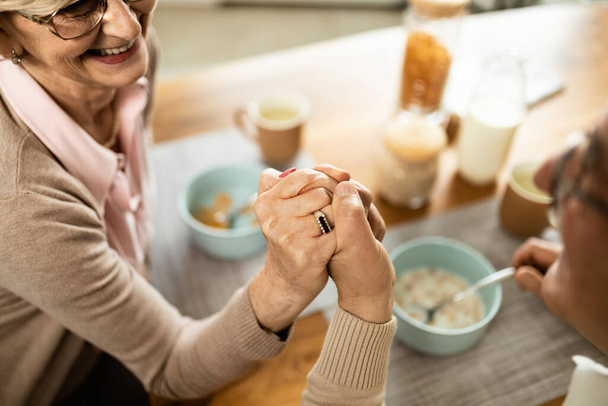 Close-up of happy mature couple holding hands and talking while having breakfast at home.  - Фото, изображение