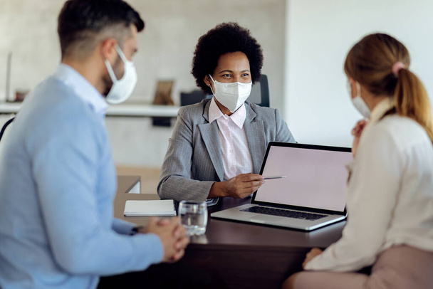 Happy black bank manager using computer while talking to a couple during the meeting in the office. All of them are wearing protective face masks due to coronavirus pandemic.  - Foto, Imagem