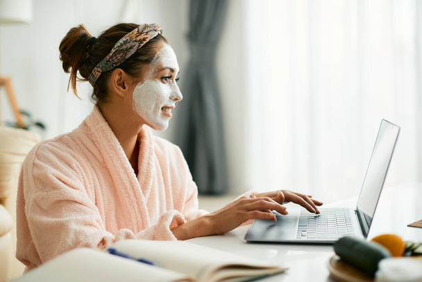 Young woman with beauty facial mask using laptop while relaxing at home. - 写真・画像
