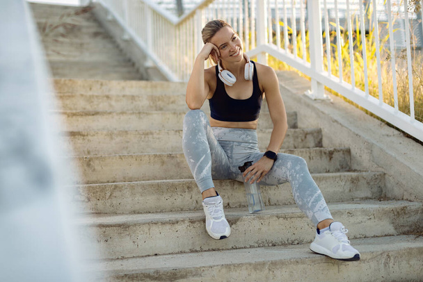 Happy athletic woman relaxing on the stairs while having water break outdoors.  - Foto, afbeelding