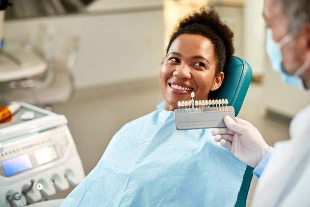 Happy African American woman and her orthodontist choosing shade of implants during dental appointment at dentist's office.  - Zdjęcie, obraz