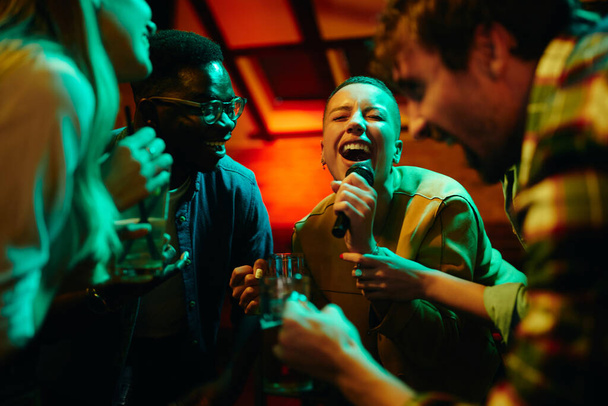 Group of carefree friends singing karaoke and having fun on night party in a bar. Focus is on black woman singing. - Zdjęcie, obraz